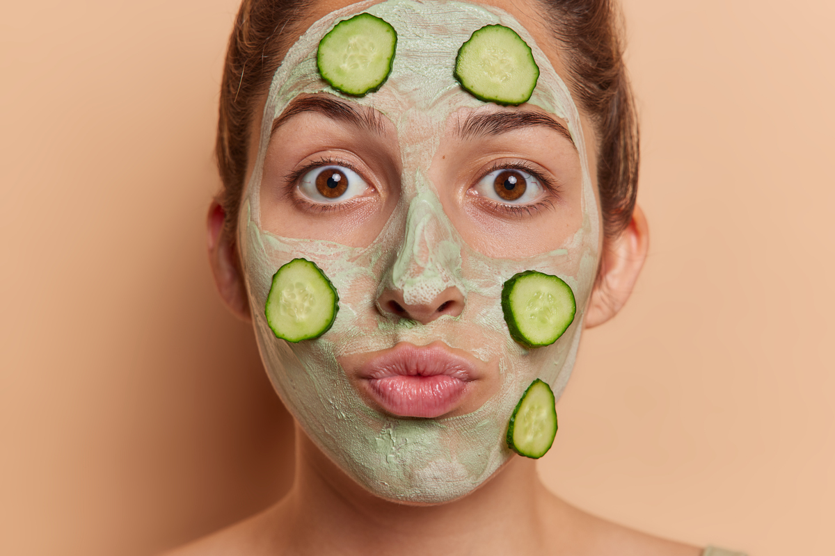 Młoda Europejka z zaokrąglonymi ustami nakłada maseczkę na twarz z plasterkami świeżego ogórka w celu odmłodzenia skóry i zmniejszenia drobnych zmarszczek